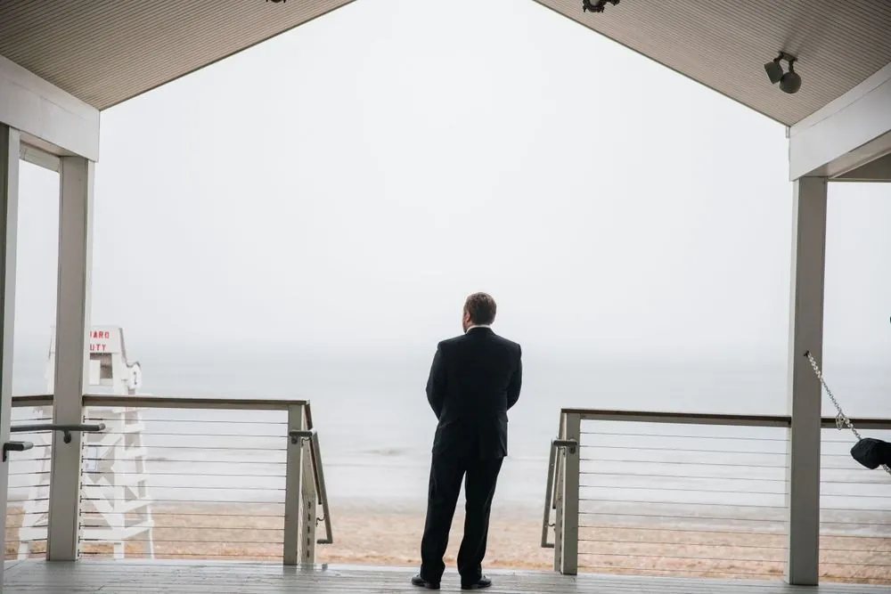 Groom waits alone under the pavilion roof facing a fogged-in beach and lifeguard chair