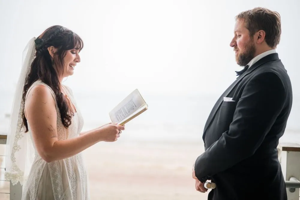 Dana reads from a small vow booklet as Michael listens with the grey beach behind him