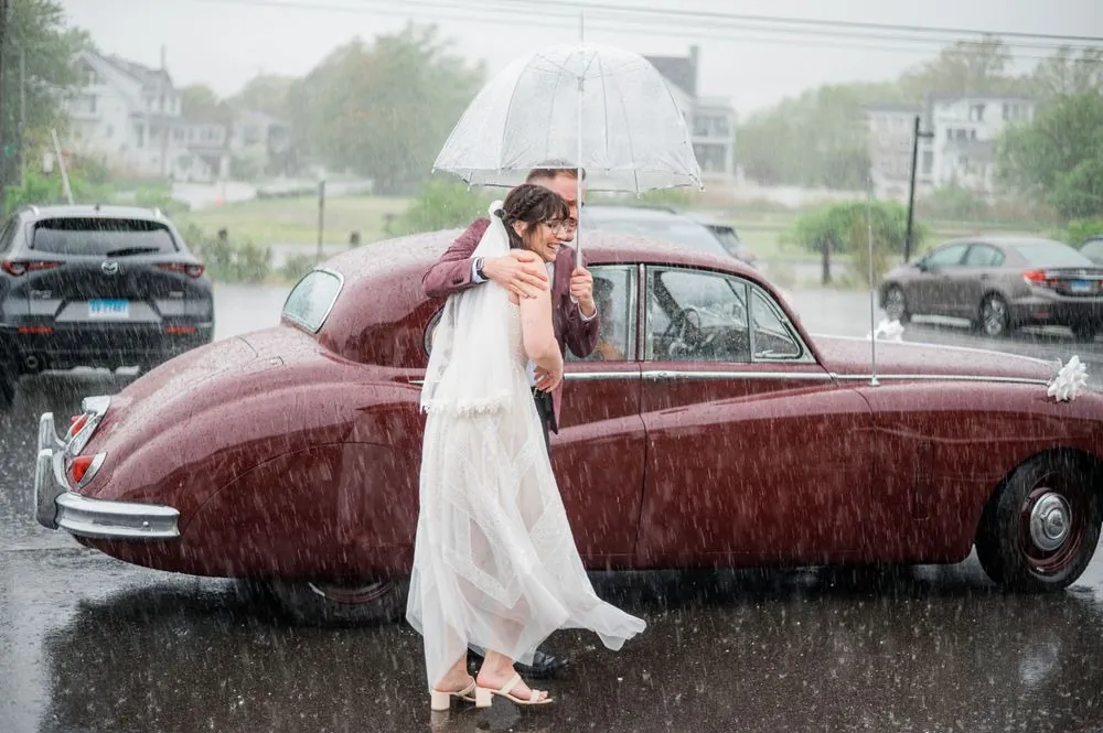 Bride laughs in a downpour beside a vintage burgundy car as a friend holds a clear umbrella
