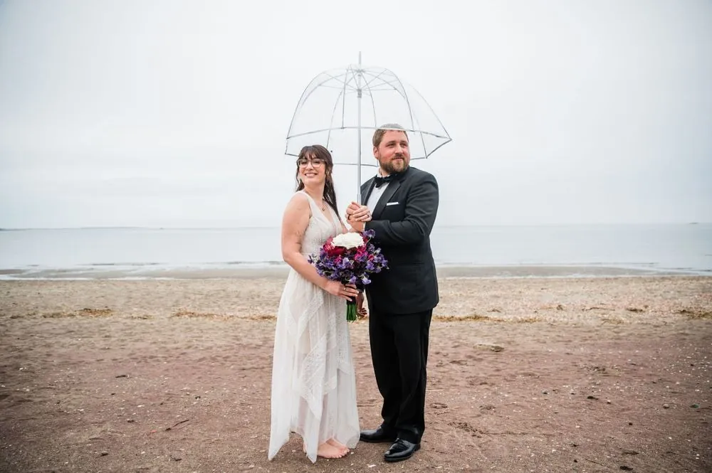 Dana and Michael share a clear umbrella on a misty beach, her purple bouquet in hand