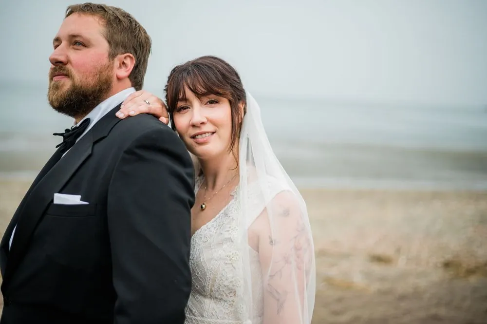 Dana leans her head on Michael's shoulder, her veil soft against the grey water