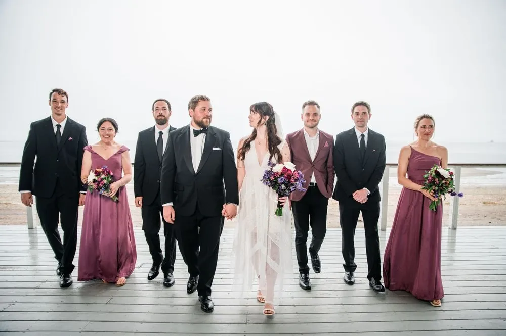 Wedding party in mauve gowns and black tuxedos walks across the wet pavilion deck