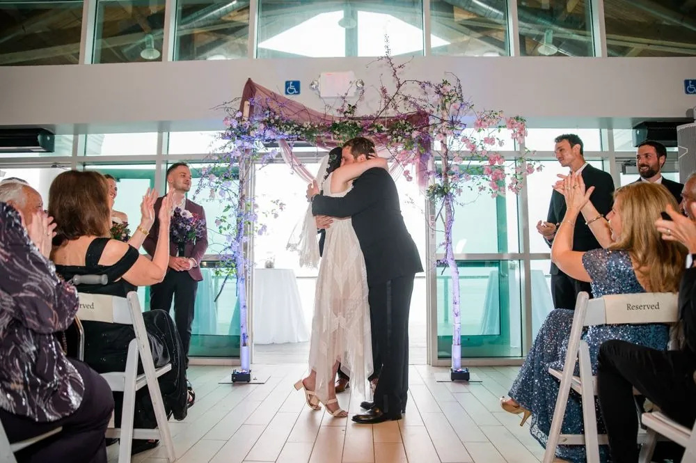 Dana and Michael embrace under a birch arch of blossoms as guests clap from white chairs
