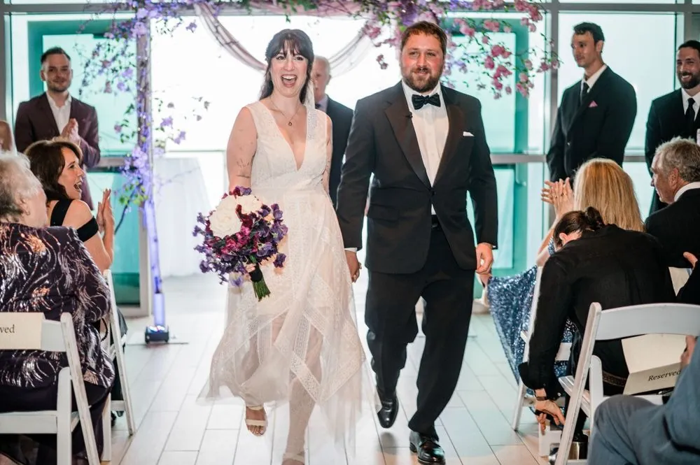 Dana and Michael walk back up the aisle laughing, her plum bouquet at her side