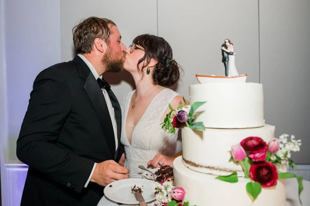 Michael kisses Dana beside a white cake trimmed with red ranunculus and tulips
