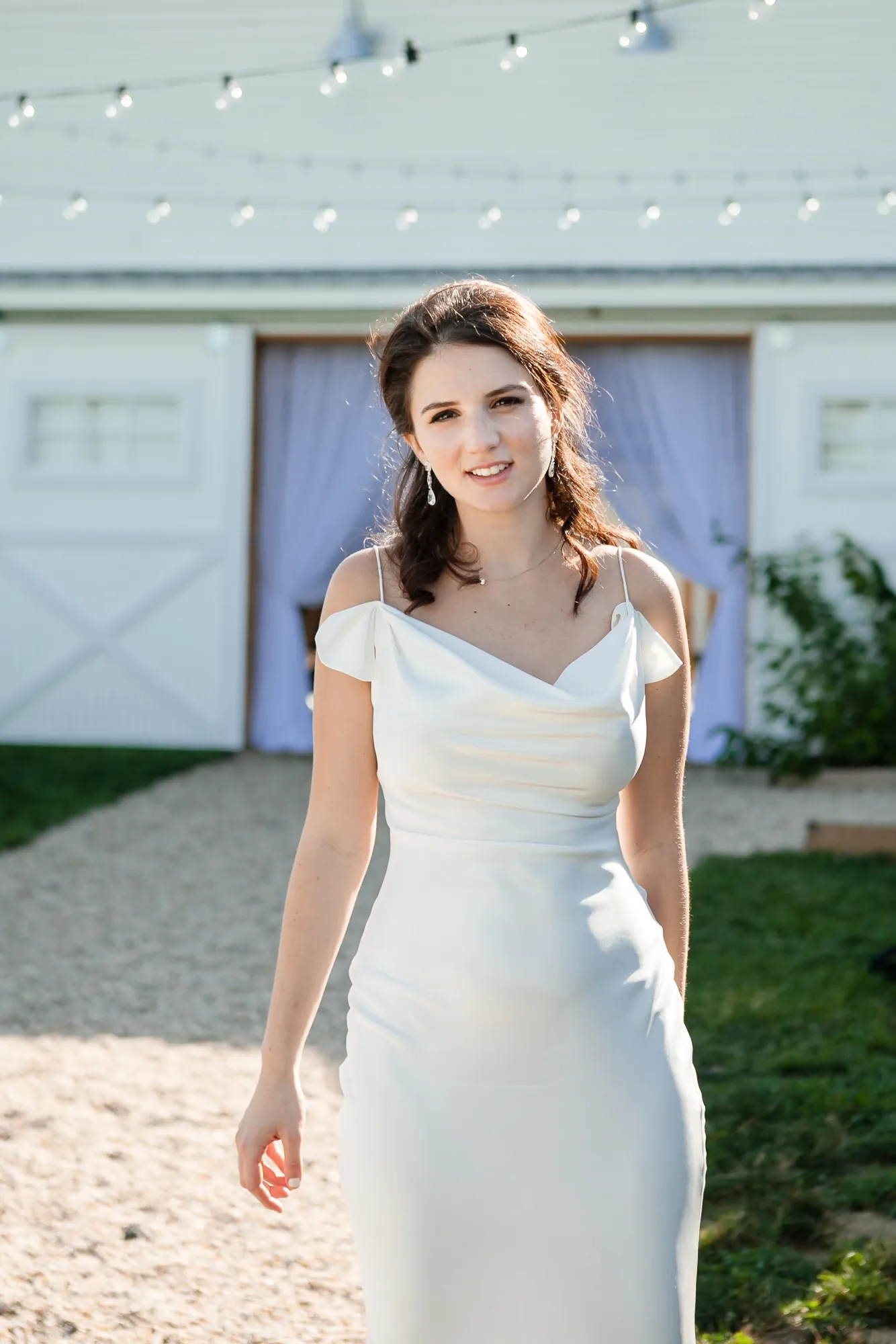 The bride in a silk slip dress steps away from the white barn doors on a sunny path