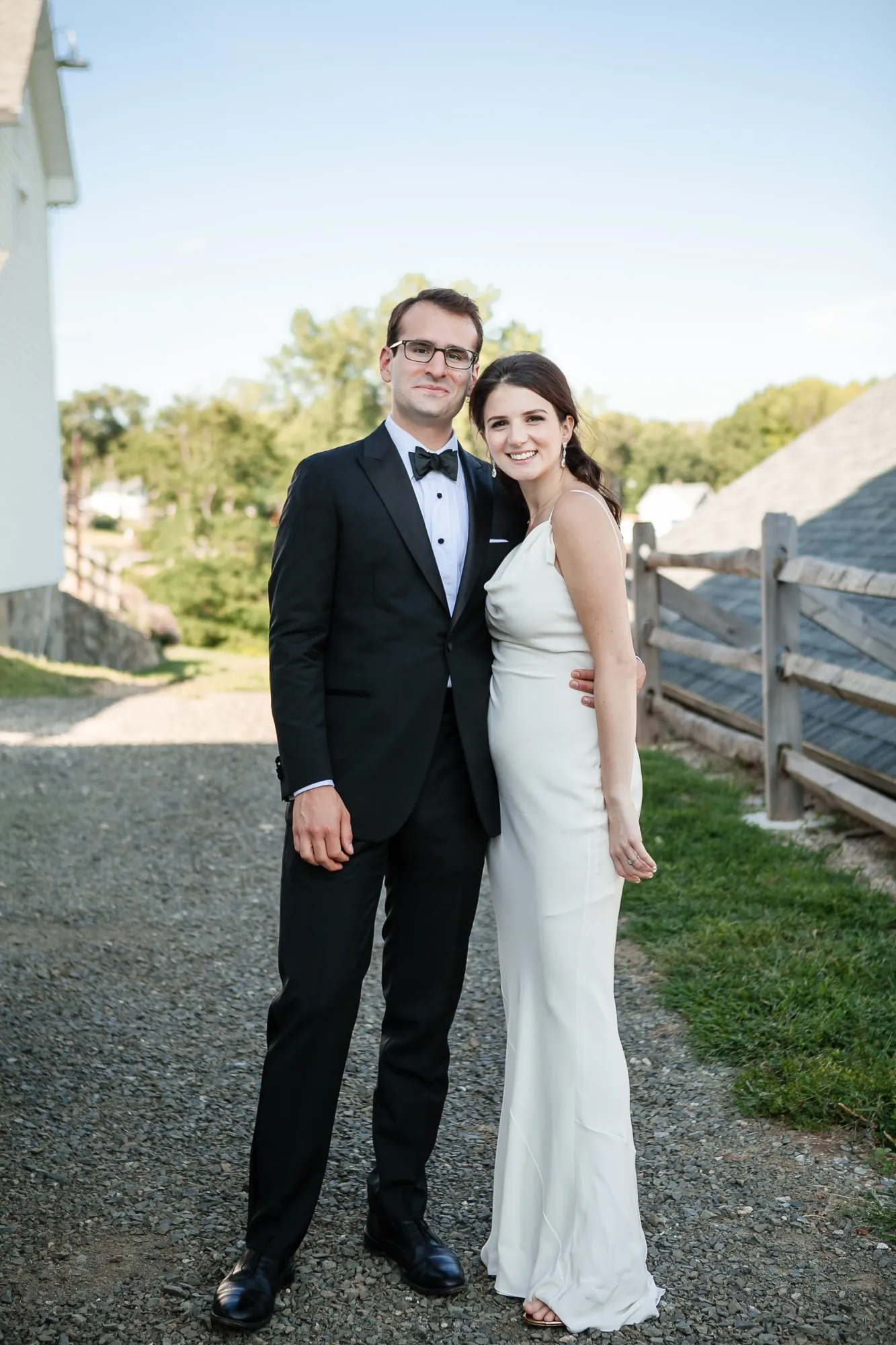 EJ and Alex stand close on a gravel path beside a white fence and barn