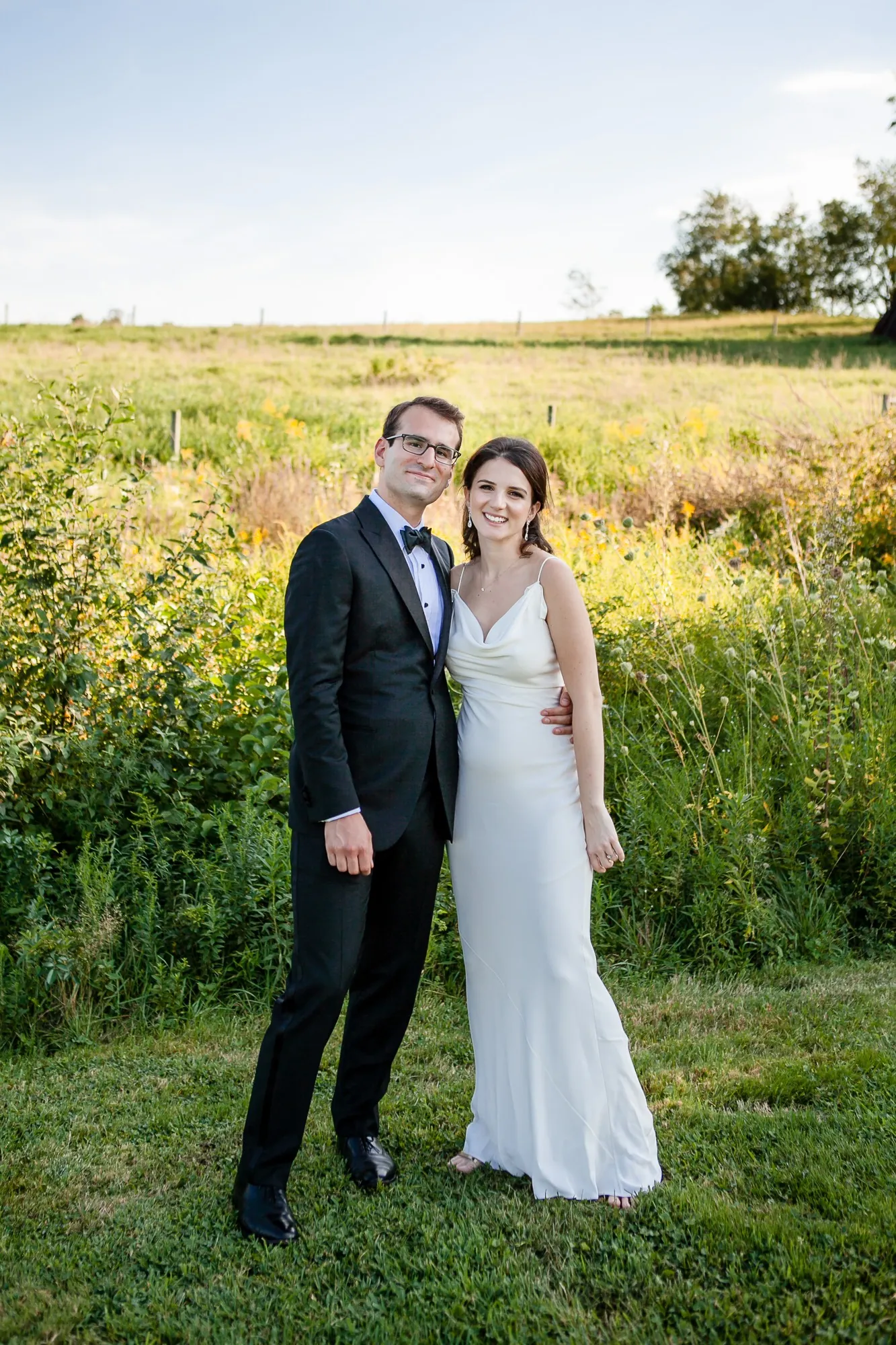 EJ and Alex stand together in tall green grass with the farm's trees behind them