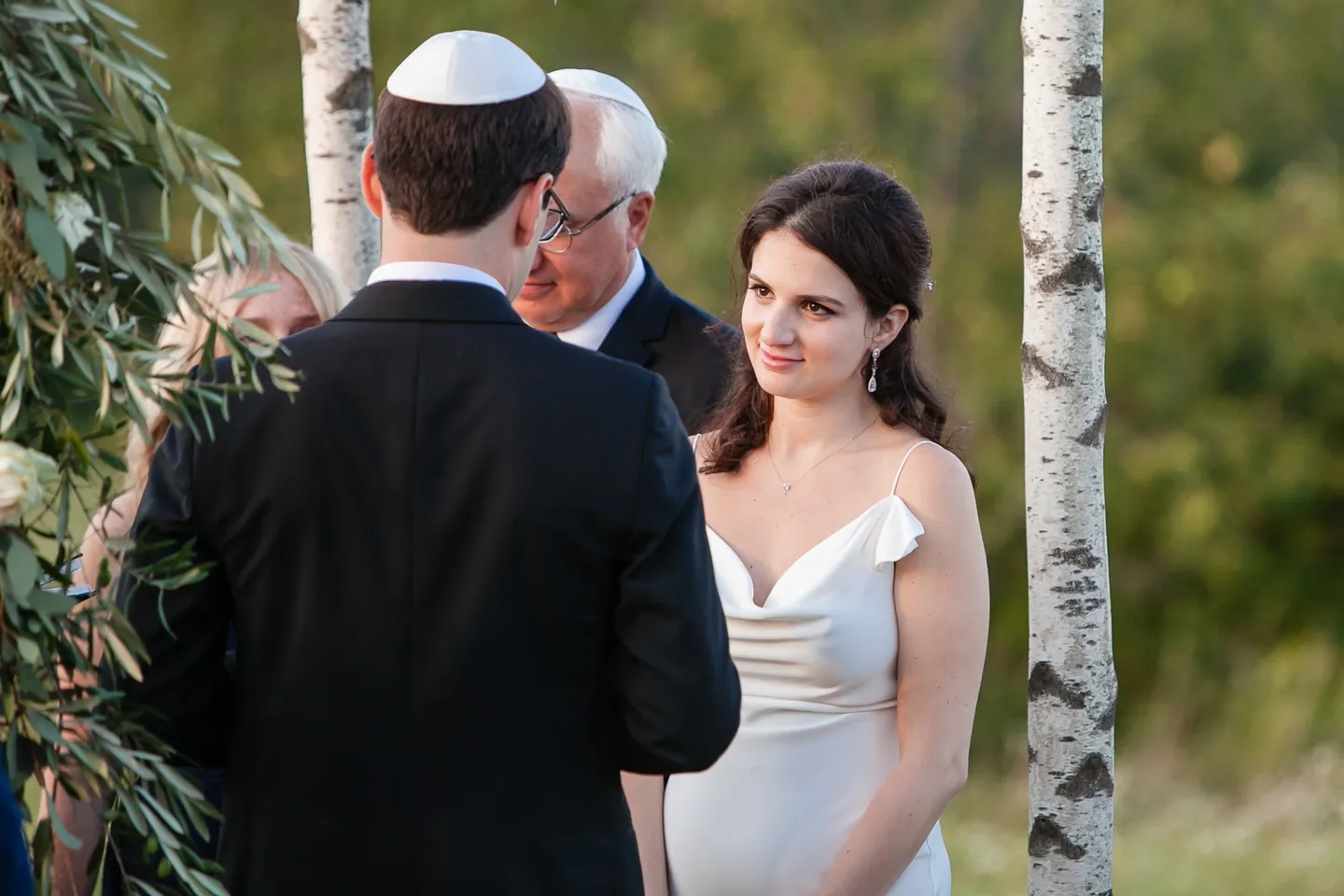 The bride smiles at the groom in his white kippah beneath a birch chuppah