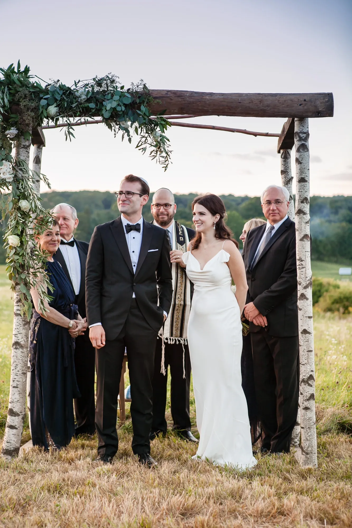 The couple stand under the birch chuppah with family on either side in the field
