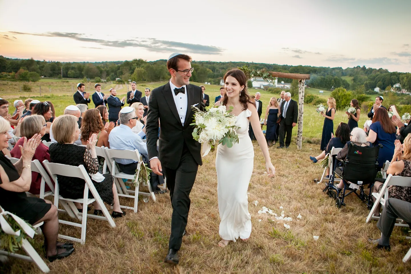 EJ and Alex walk back up the aisle past clapping guests in the open field