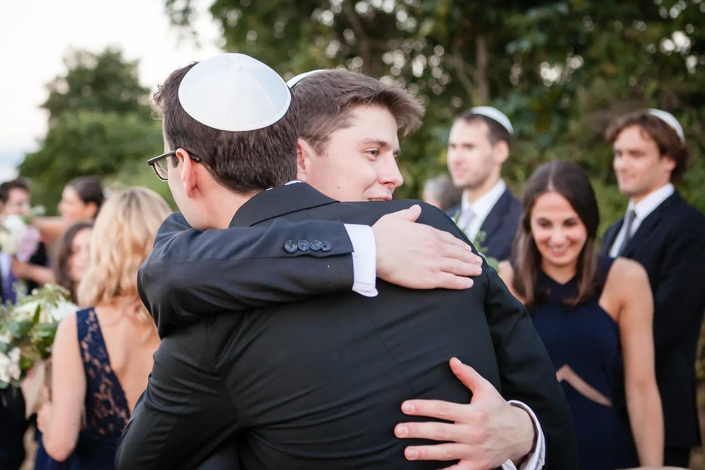 A man in a white kippah hugs another guest tightly after the ceremony as others smile