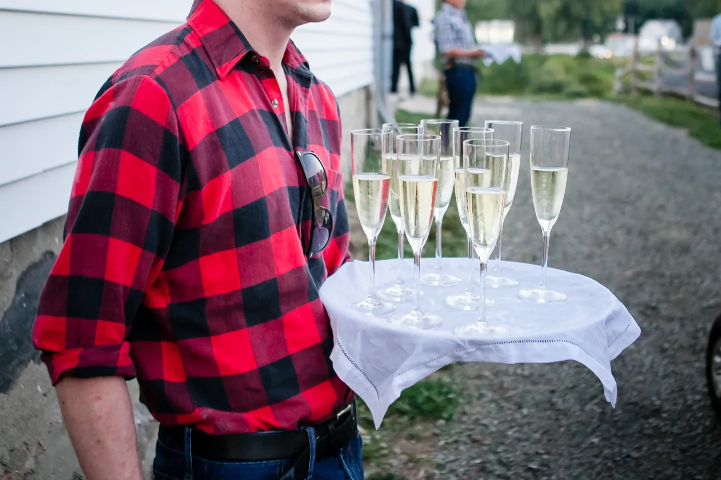 A man in red flannel stands beside a table of champagne flutes on the gravel