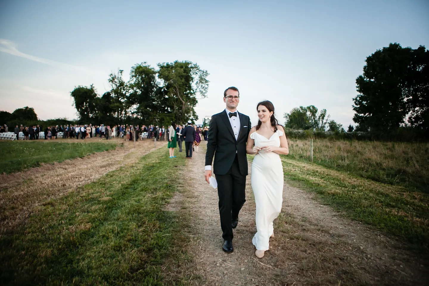 EJ and Alex walk a dirt farm road smiling, guests trailing behind them at dusk