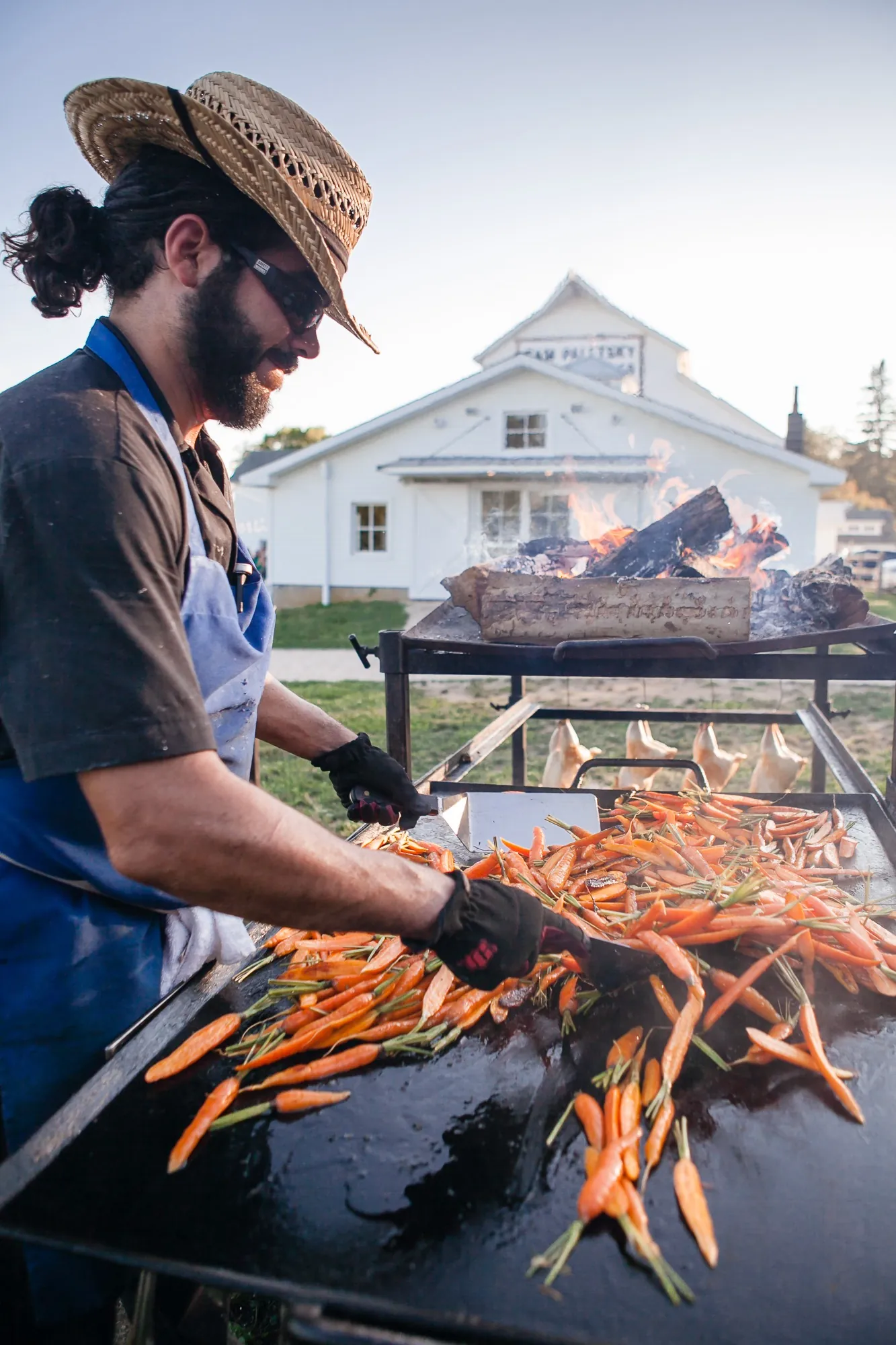 A cook in a wide-brimmed hat turns whole carrots on an open grill by the farmhouse