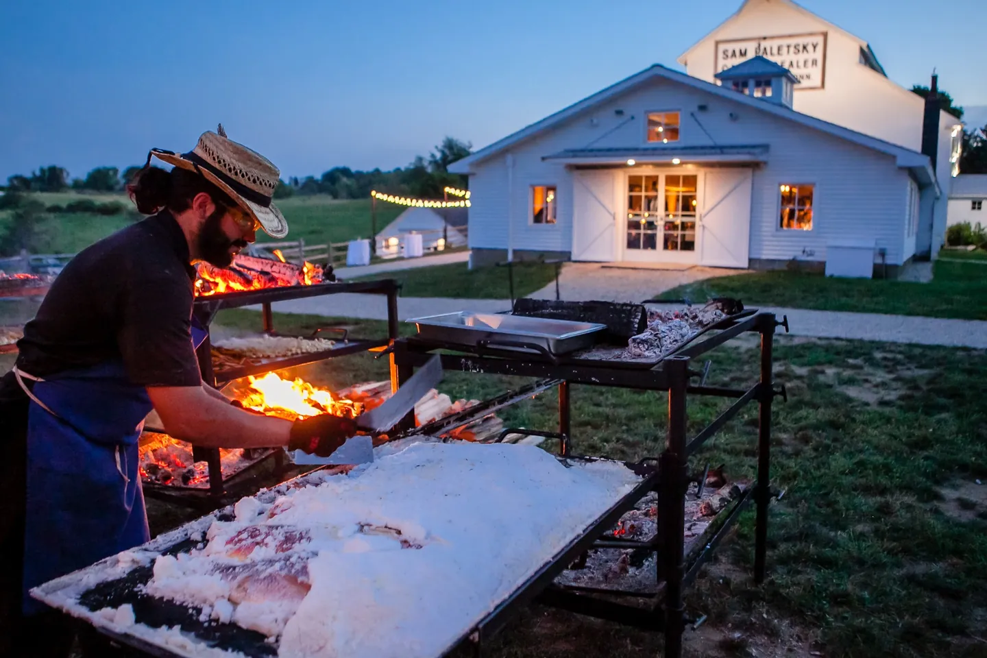 A cook tends a large open fire at dusk beside the lit white barn