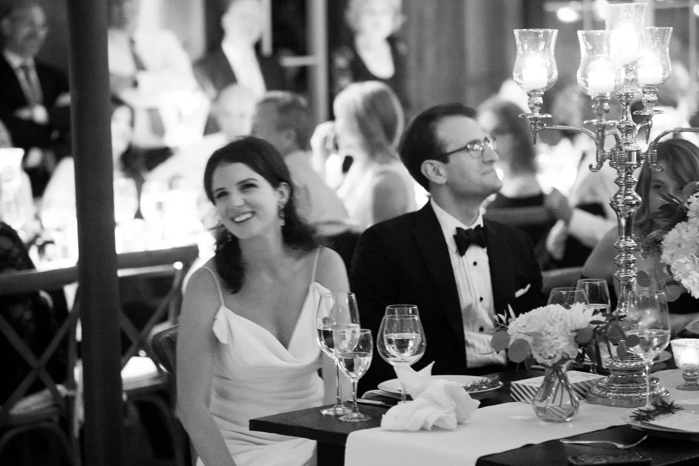 The bride laughs at the dinner table beside the groom, in black and white