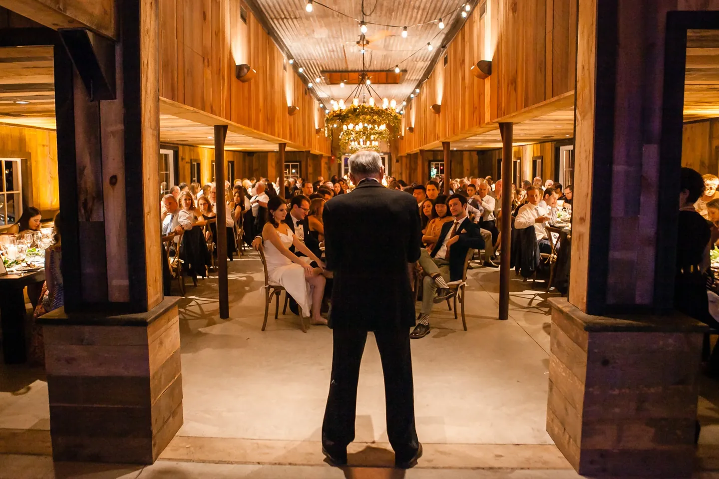 A guest gives a toast at the end of the long barn as the tables listen