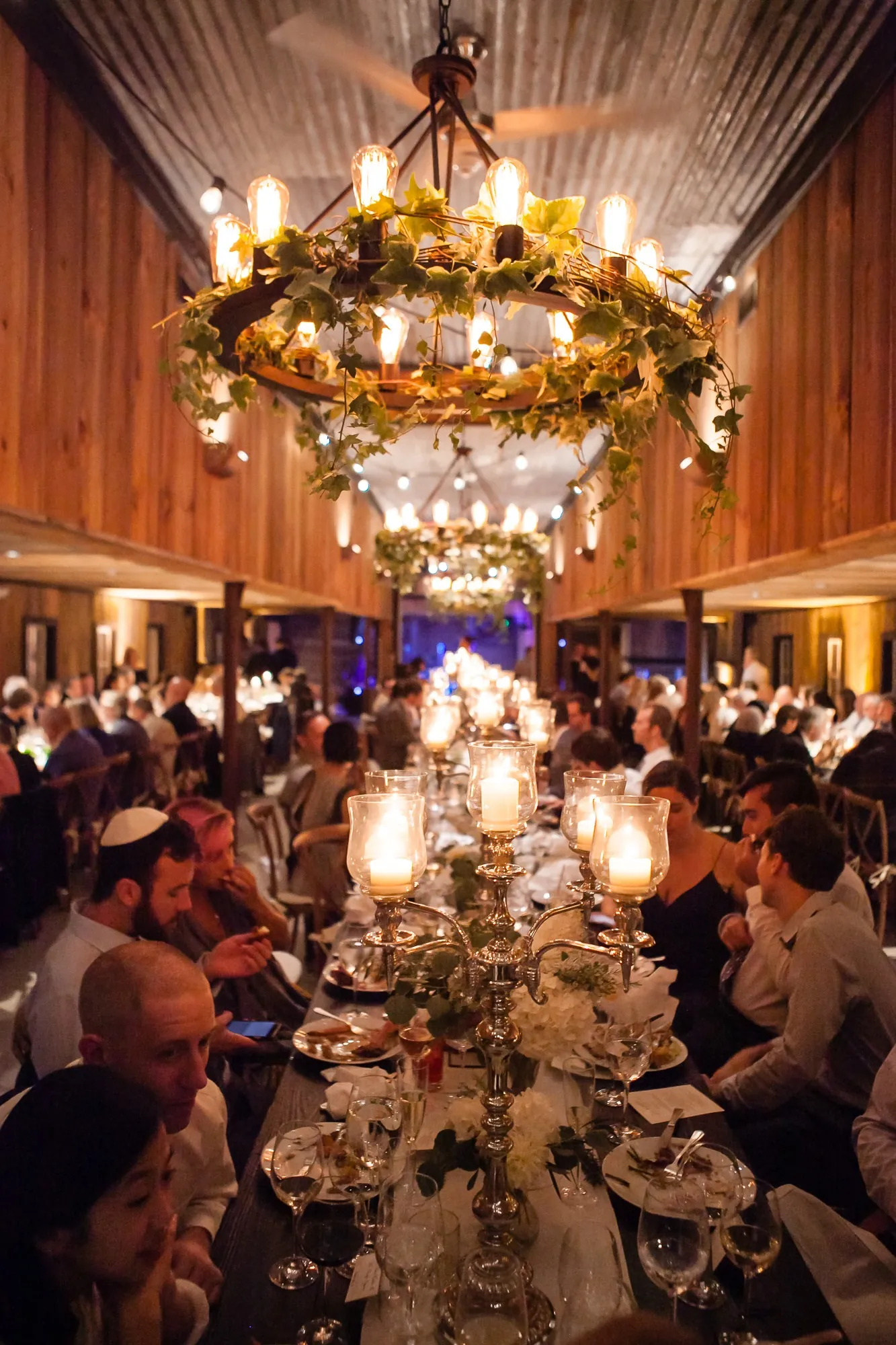 Guests dine by candlelight under a chandelier wound with greenery in the barn
