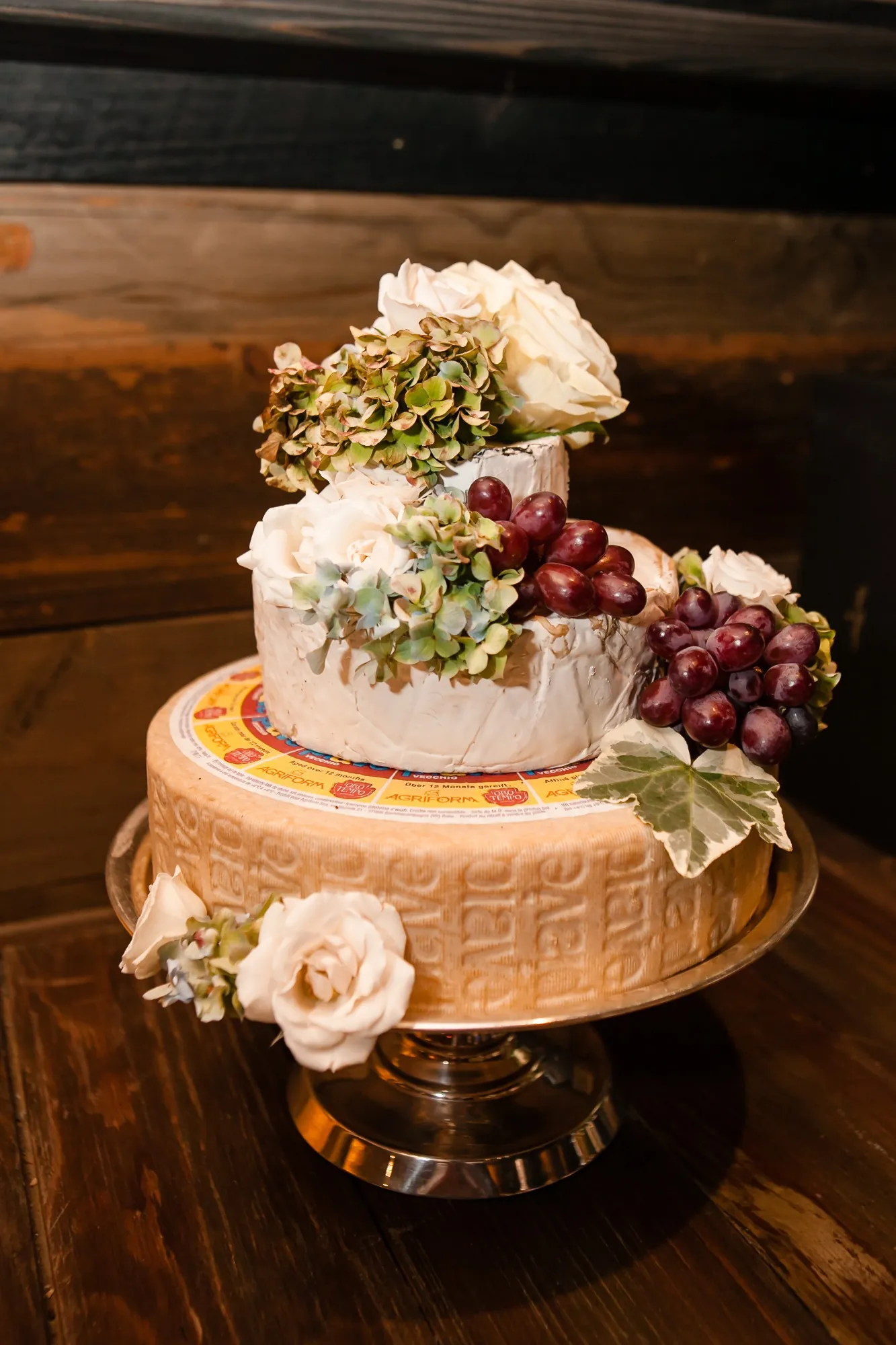 A tiered cake decorated with grapes and flowers sits on a wooden slab