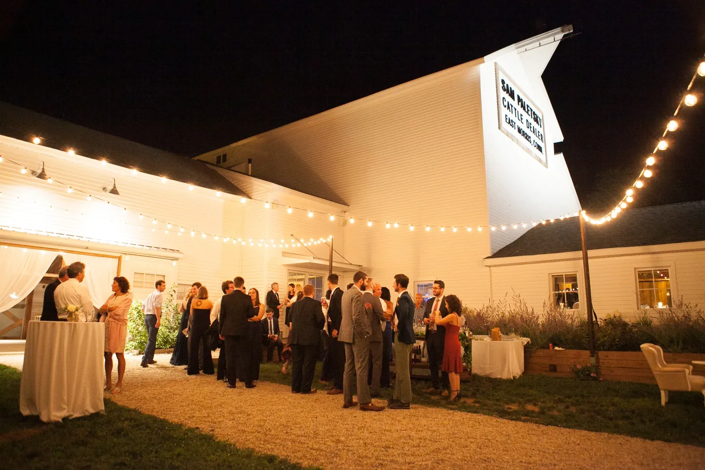 Guests gather under string lights outside the white barn after dark