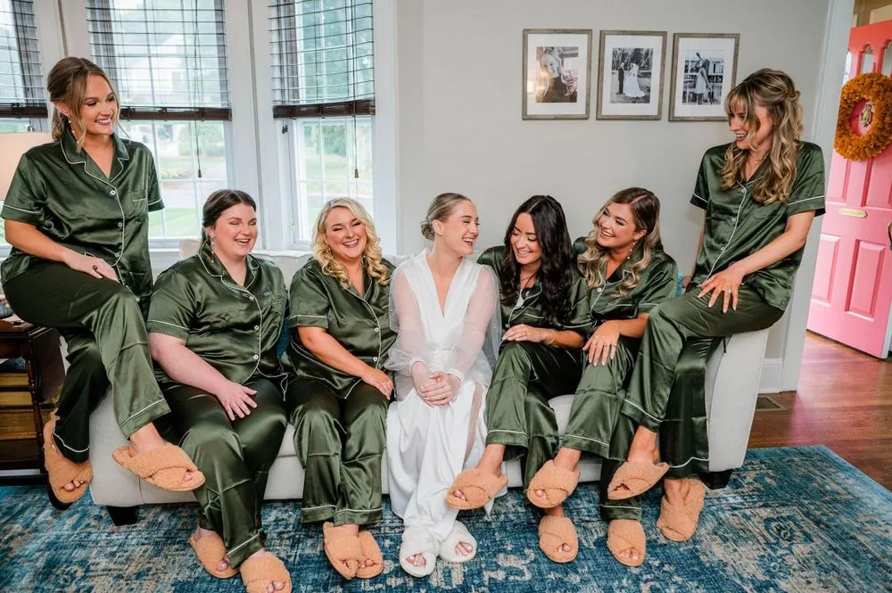 Bridesmaids in green satin pajamas sit on a couch around the bride in a white robe