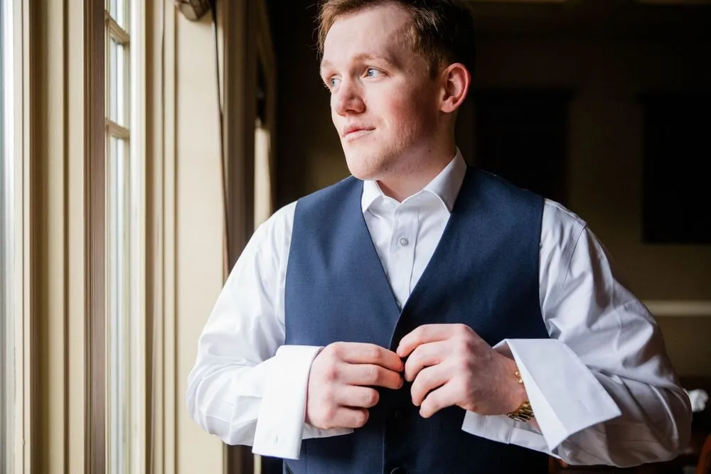 The groom buttons his navy vest beside a tall window in soft side light