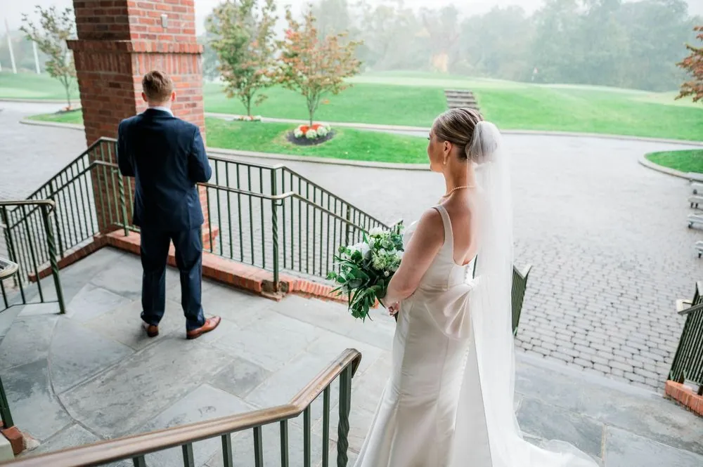 The bride walks up behind the groom on the stone steps above the foggy golf course