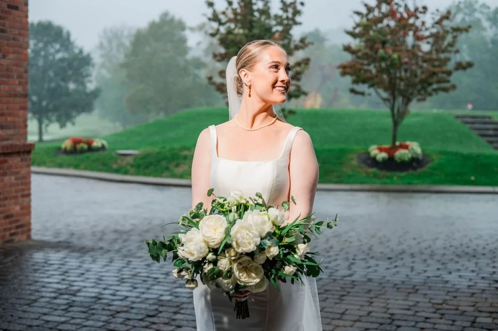 The bride holds a white rose bouquet on the brick terrace with fog over the course