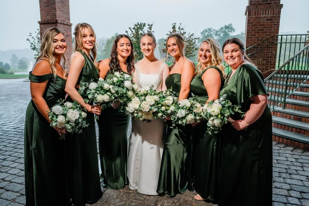The bride stands with her bridesmaids in emerald satin gowns on the brick terrace