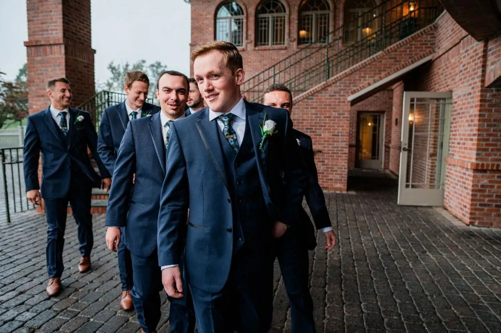 The groom and groomsmen in navy suits walk together across the brick patio