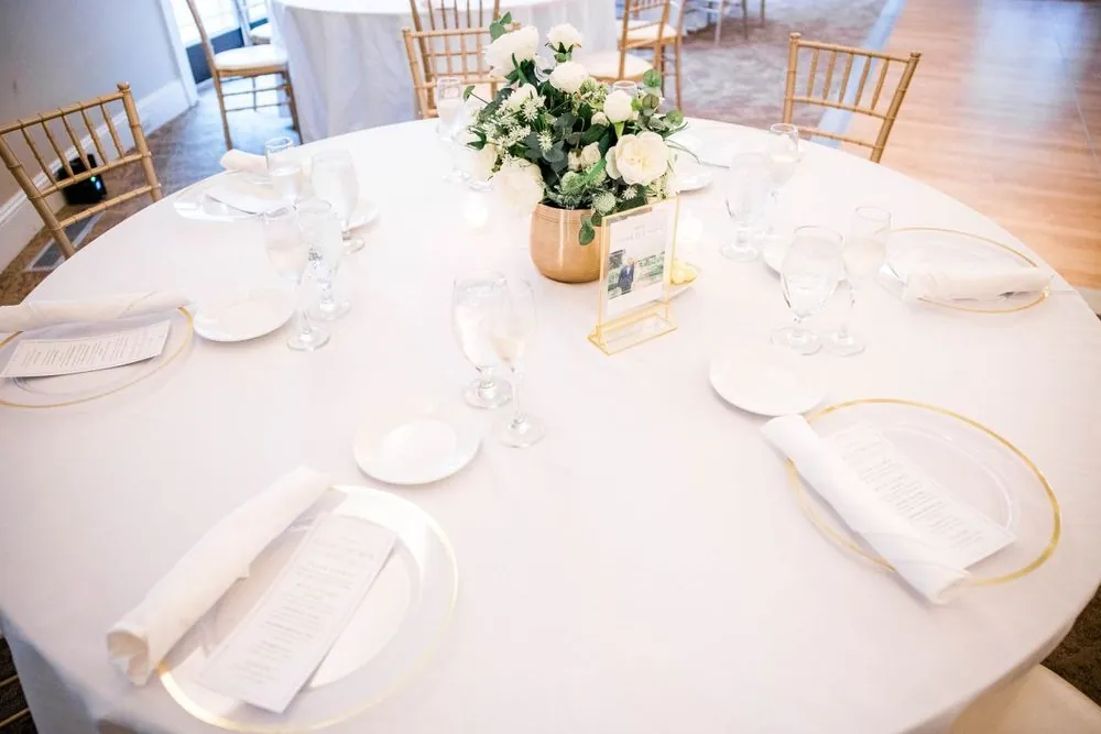 A round reception table set with white linens, gold chargers and a low white centerpiece