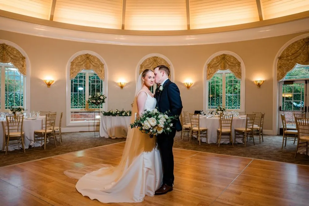 Elizabeth and Colsen kiss in the center of the empty ballroom beneath tall arched windows