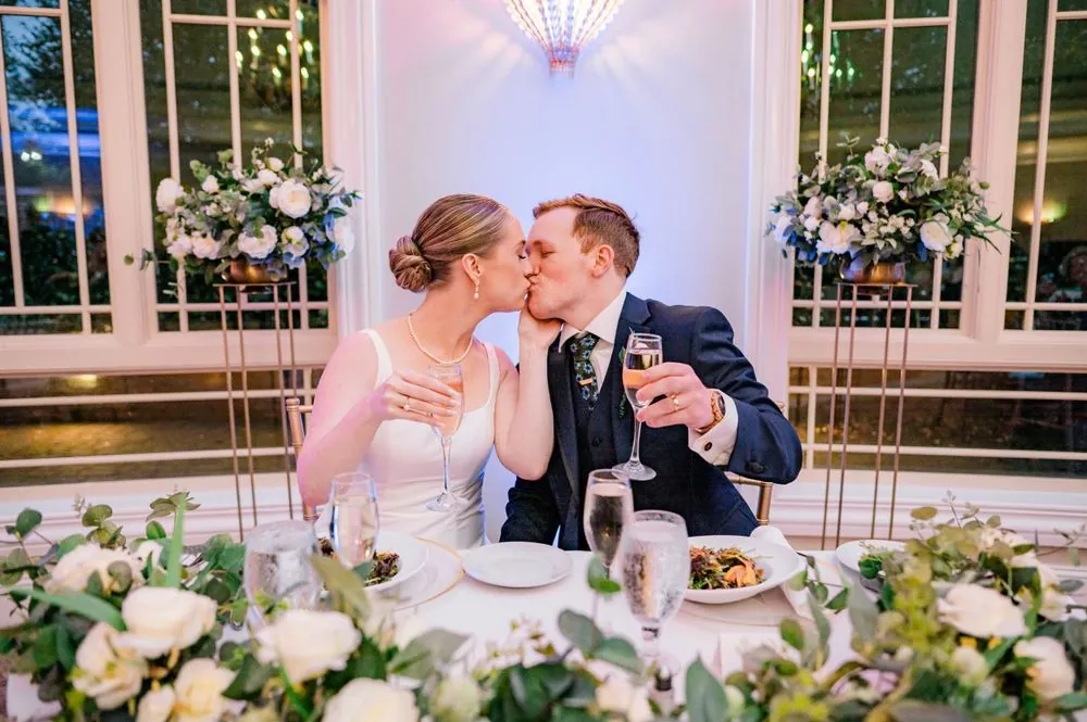 The couple kisses at a sweetheart table covered in white flowers and greenery
