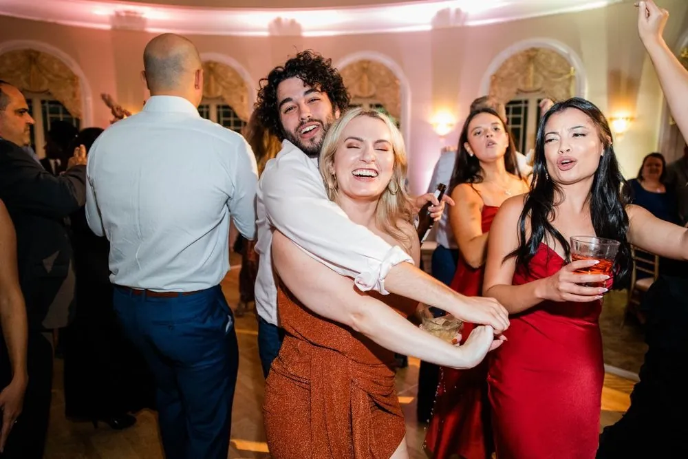 Guests in bright dresses sing and dance together under pink ballroom lights