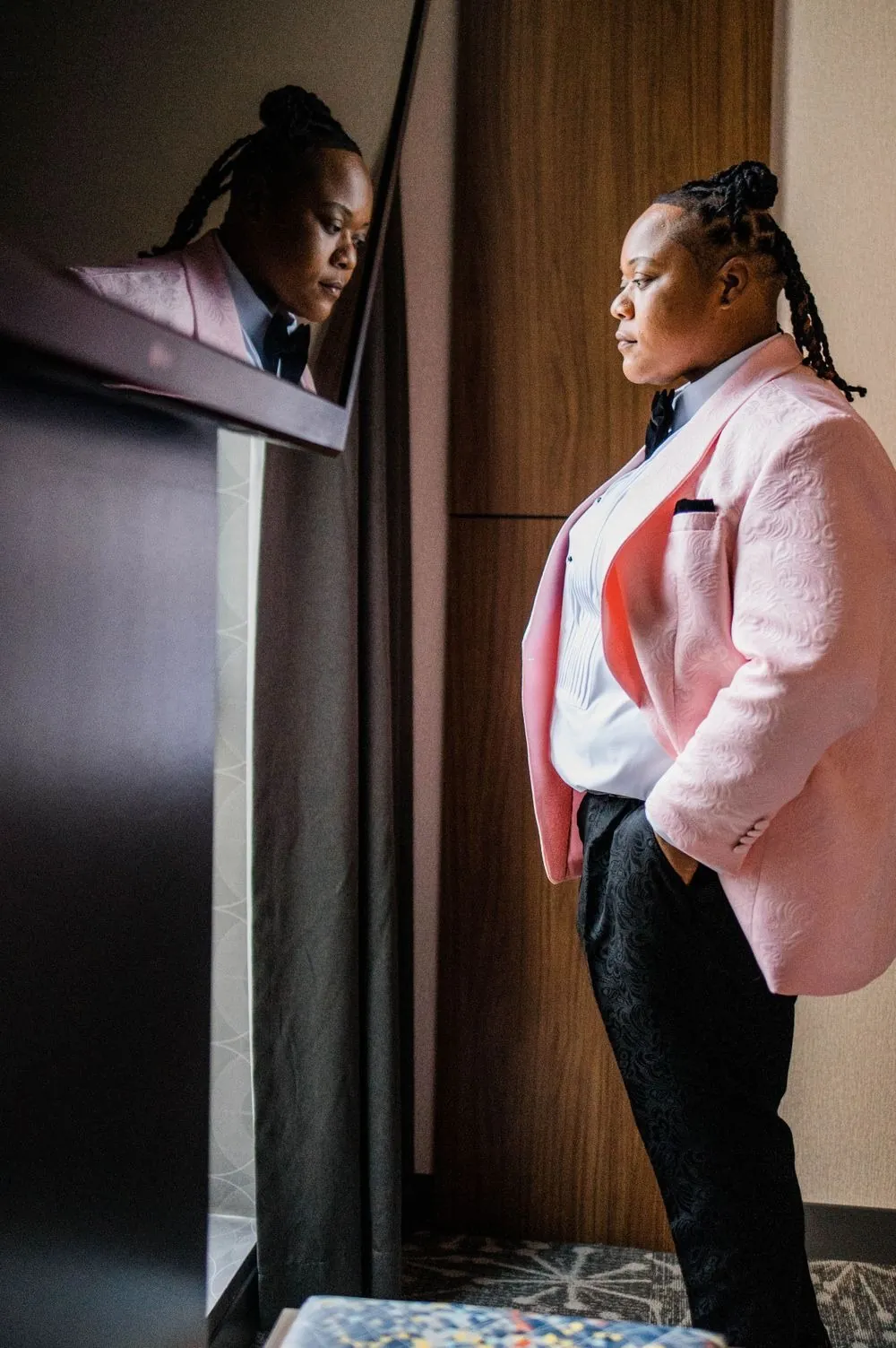 Dressed in a blush pink tuxedo jacket, one of the couple looks out a tall window
