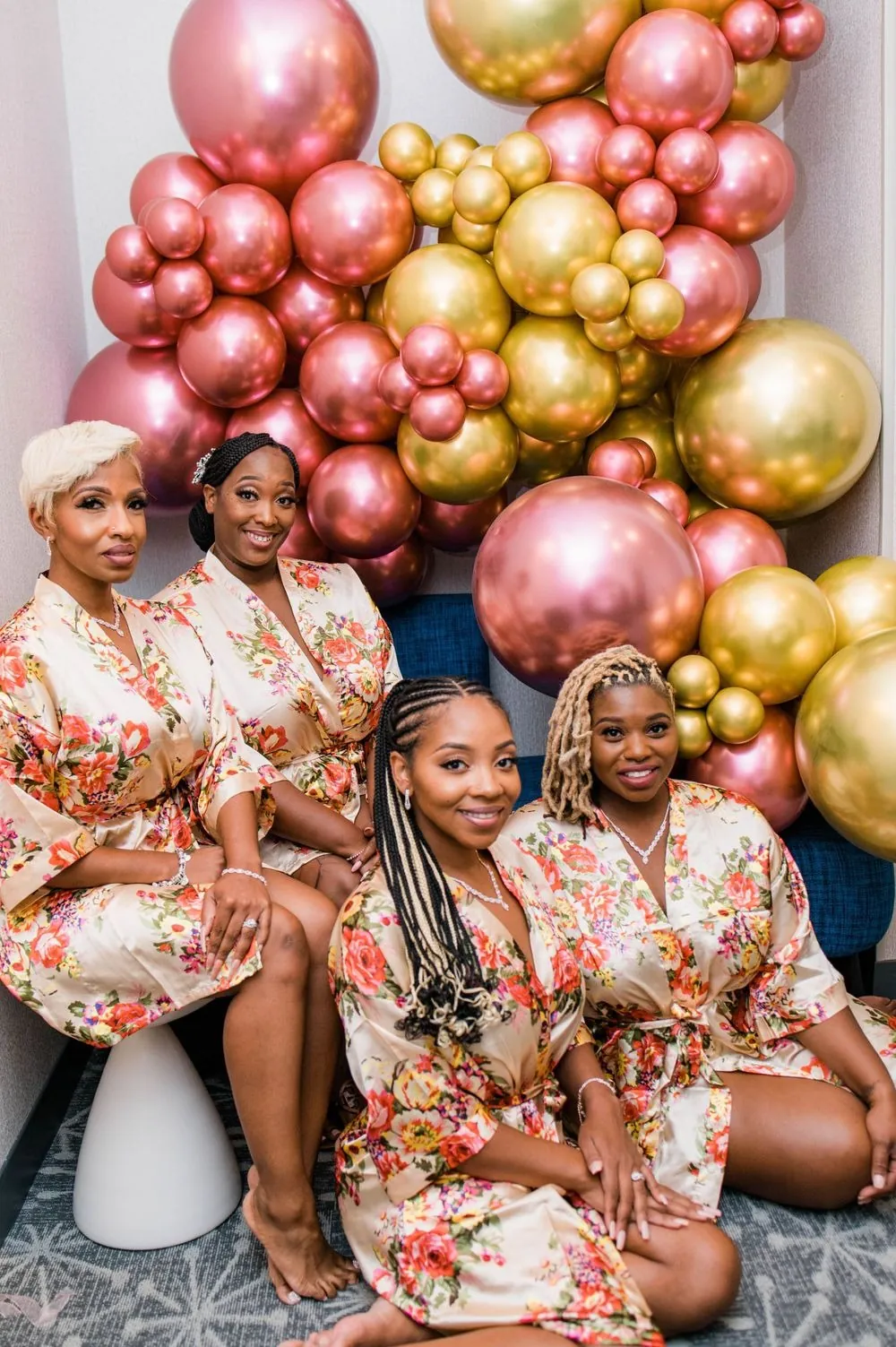Bridesmaids in floral robes pose in front of a wall of pink and gold balloons