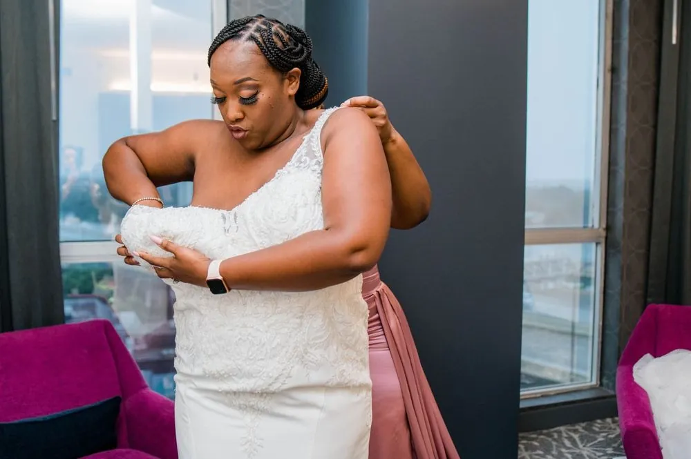 The bride adjusts her white gown beside floor-to-ceiling city windows