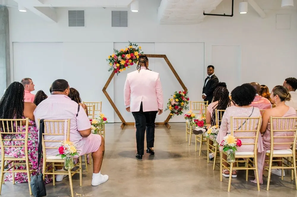 Guests on gold chairs face a hexagon arch of flowers in the bright ceremony room