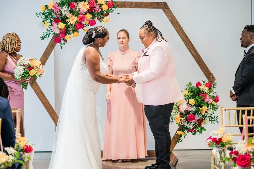 The couple faces each other under the flower arch with bridesmaids in blush beside them