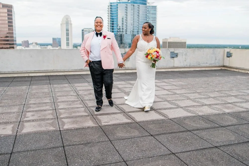 The couple walks hand in hand across the rooftop with towers behind them