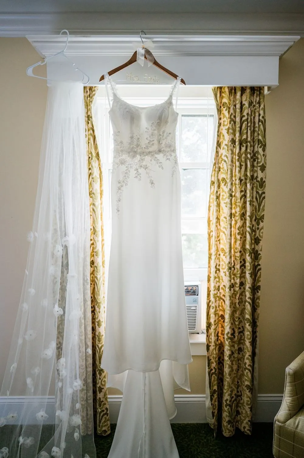 The wedding gown hanging in a window framed by yellow patterned curtains
