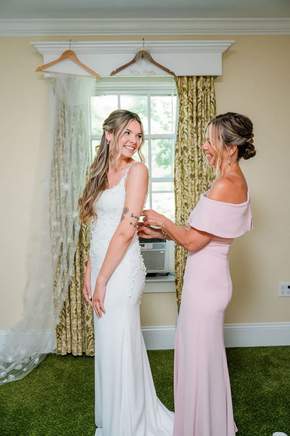 A bridesmaid in blush helps the bride with her gown by the window