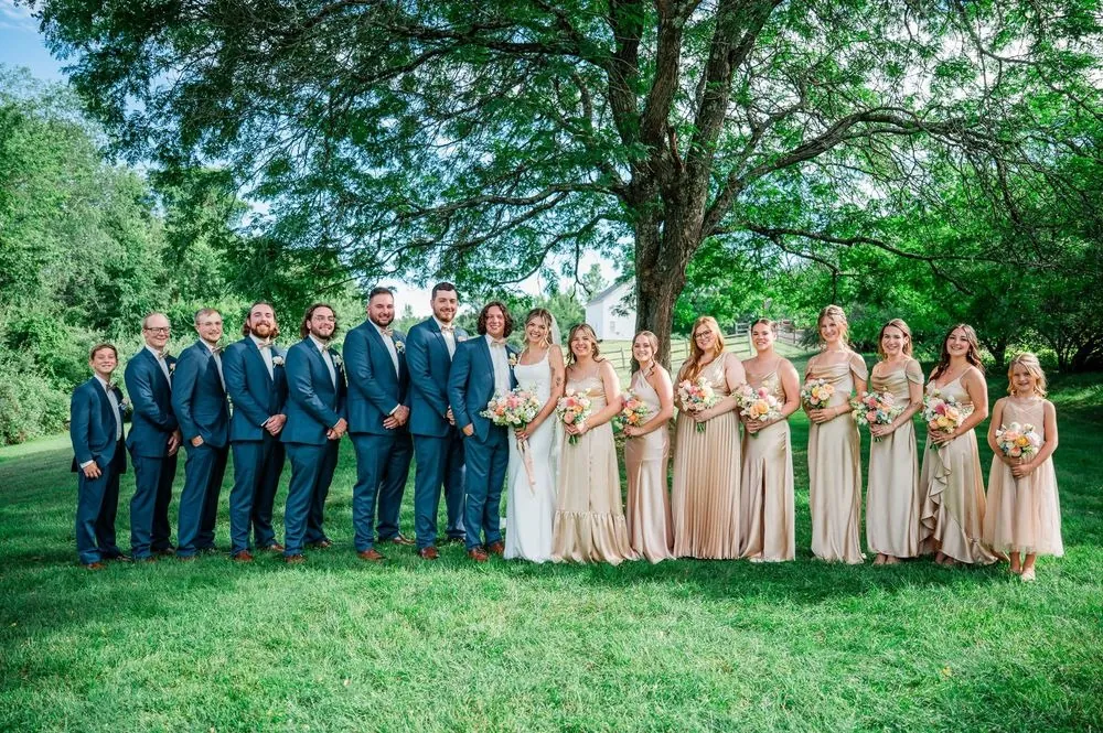 The full wedding party in blue suits and champagne gowns on the lawn
