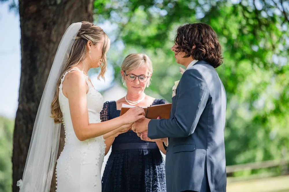 The couple exchanges rings beneath a big tree as the officiant holds a leather notebook