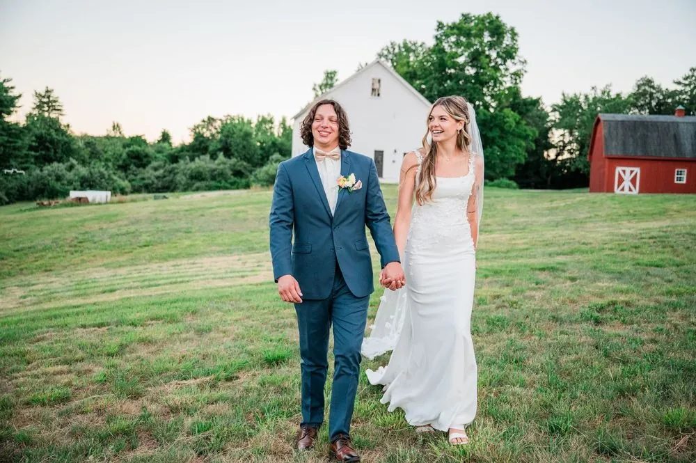 Fallon and Dom walk hand in hand across a field with white and red barns behind