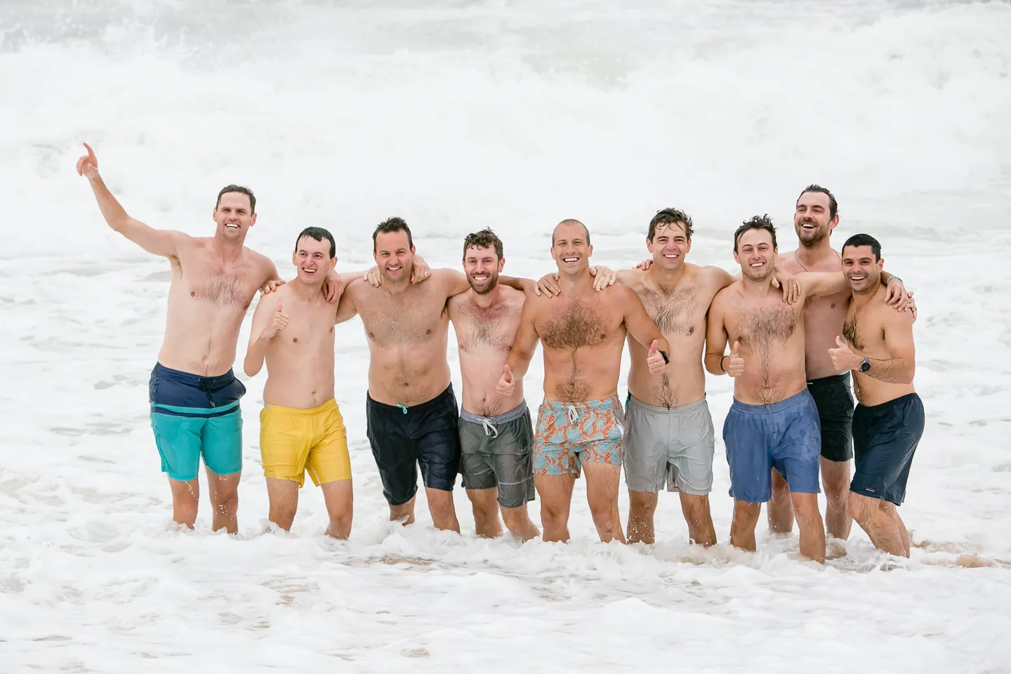 Groomsmen in bright swim trunks stand waist deep in foamy surf, arms raised, under a grey sky