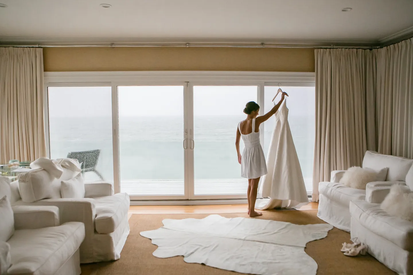 The bride reaches for her gown hanging in front of wide windows looking out to the ocean