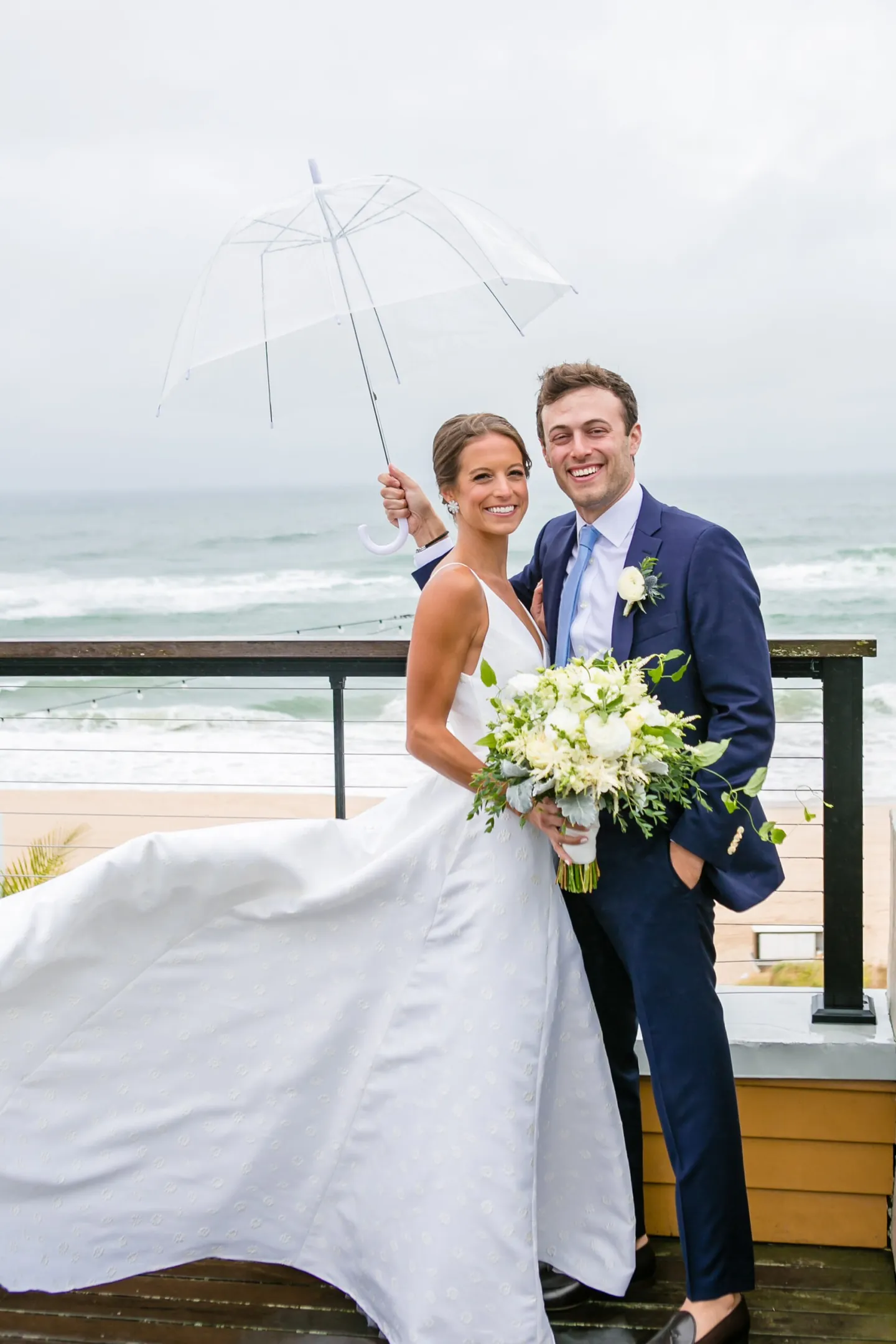 Hannah and Mark smile under a clear umbrella on a deck above the beach, her train blowing sideways