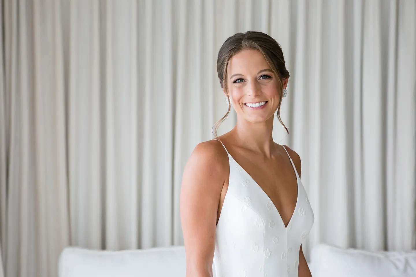 The bride smiles in a plunging white gown against soft grey curtains